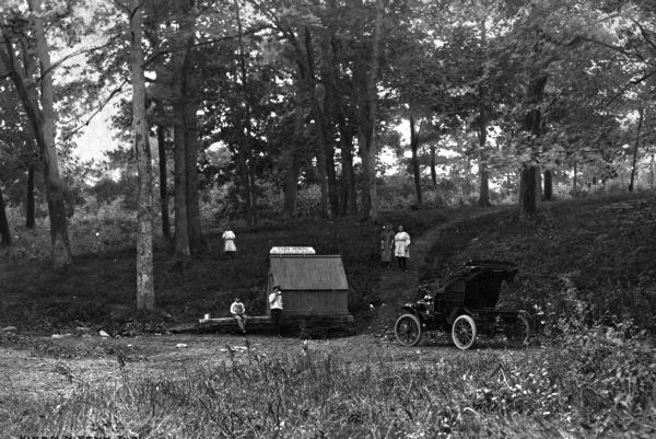 Travelers stop by a spring in the woods for water with their early-model automobile. Two boys are standing at the spring, and a young girl stands above them on the left on the side of the hill covered with trees. Above the boys on the right are two girls are standing on a trail. A sign on top of the building at the spring reads: "Do not Deface this building."