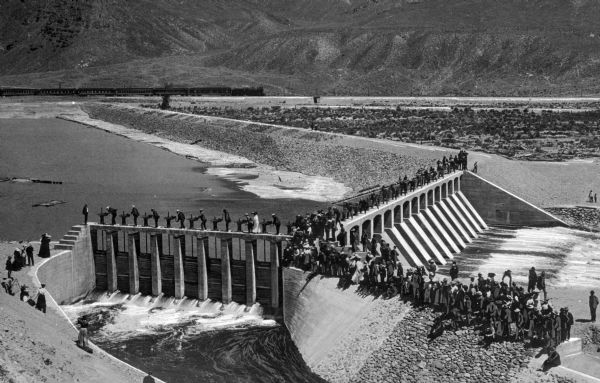 Elevated view of Derby Diversion Dam with a crowd gathered on the rim of the dam and individuals standing along its top. A train cuts across the horizon in the distance and mountains rise in the background.
