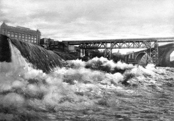 View of the roiling lower falls on Spokane River with two bridges, and a large stone building with a sign that reads: "Washington Water Power Co." is above the falls in the background on the left.
