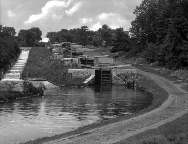 Five combined locks between the Hudson River and Lake Champlain in a wooded area.  A dirt road runs along one side and a stepped concrete stream-bed runs along the other.