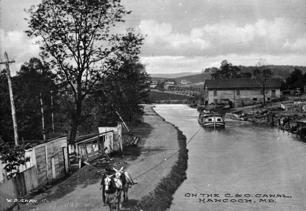Elevated view of a man with two mules on the left pulling a boat up the Chesapeake and Ohio Canal. A large wooden building is on the right bank in the behind the boat, and a hillside with dwellings and fields is in the background. Text at bottom reads: "On the C. & O. Canal, Hancock, MD."