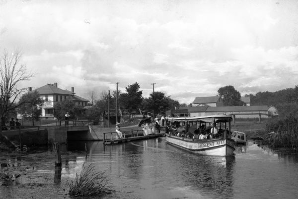 Passengers board the excursion boat "Crescent No. 1" as others look on from bridges. There is a large building in the background on the left, and a long, wooden building on the right.