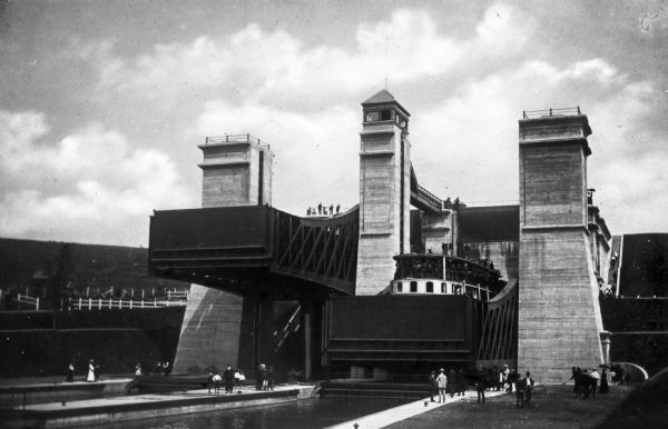 A boat entering a hydraulic lift lock located in Peterborough, Ontario, Canada. The lock structure is an imposing series of steel and concrete towers with its upper portion at a full elevation of 66 feet. Onlookers stand at the foot of and on top of the lock.