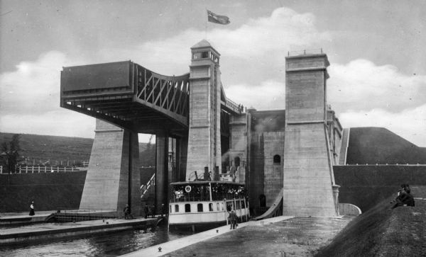 A boat entering a hydraulic lift lock located in Peterborough, Ontario, Canada. The lock structure is an imposing series of steel and concrete towers, with a flag flying on top of the tower, and is at a full elevation of 66 feet.