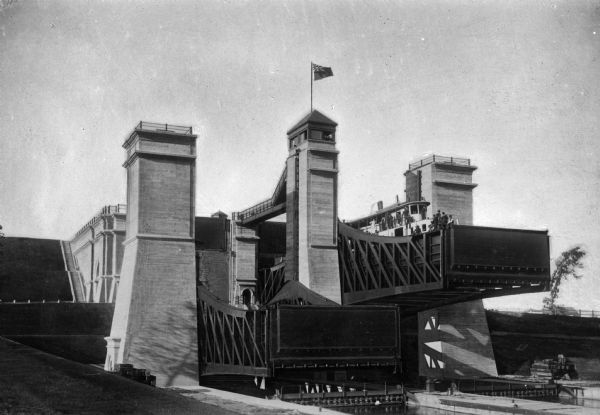 A boat half way up a hydraulic lift lock located in Peterborough, Ontario, Canada. The lock structure is an imposing series of steel and concrete towers, with a flag flying a flag at the top of the tower. The upper portion is at a full elevation of 66 feet.