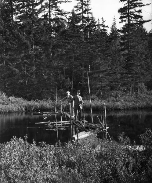 Two men in suits crossing a rickety floating bridge on Squash Pond near Hotel Glennmore.