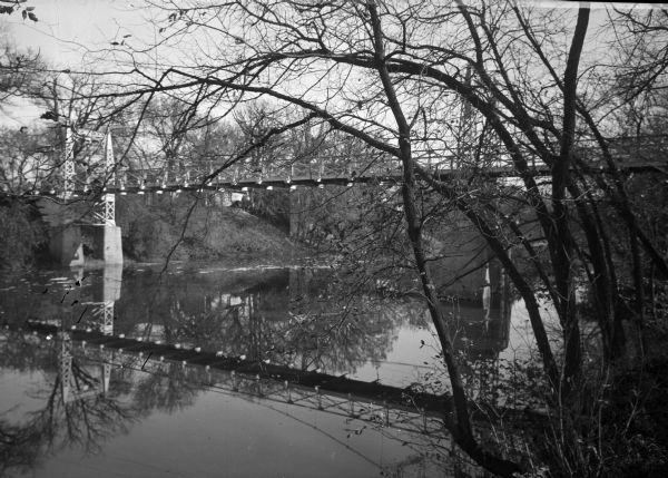 View from waterline of riverbank through a curving stand of small trees toward a footbridge on the right crossing over the Sheyenne River.