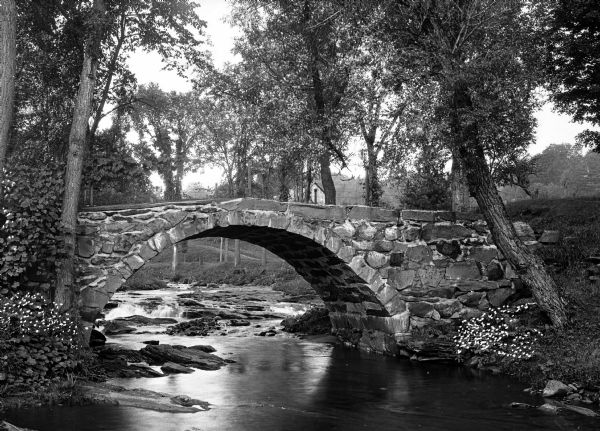 View toward a stone bridge with arch over a stream bordered with flowers and trees on South Woodstock Road.