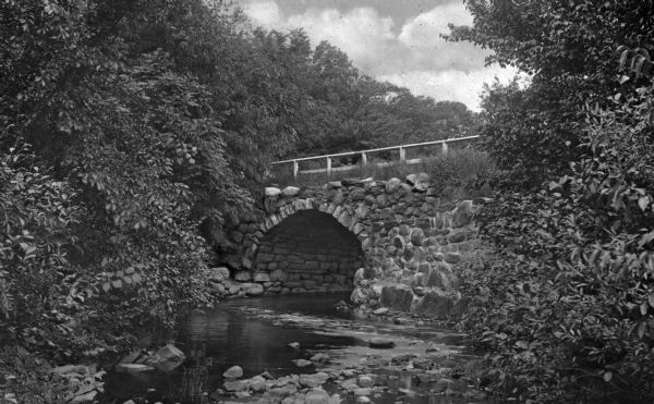 View along quiet stream lined with trees and foliage toward an arched stone bridge with a guardrail.