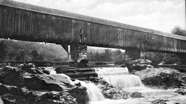 A long covered bridge and a small water fall beneath it.
