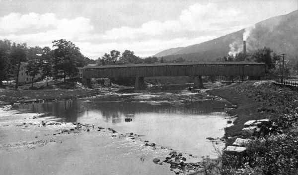 A view of a covered wooden bridge over the Juniata River with smokestacks in the background.