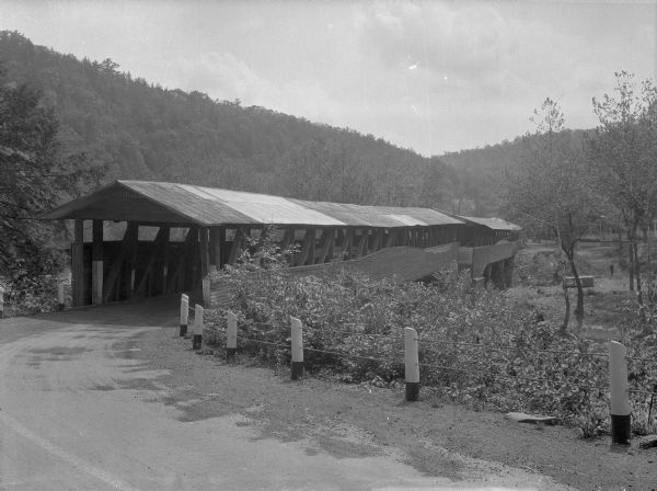 A view of a covered bridge from the road.  Forests cover the hillside in the background.