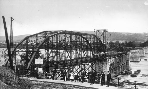 A view of the first span of the Berwick-Nescopeck bridge.  Onlookers pose for the photograph in front of a fence and completed pilings stretch across the river in the background.