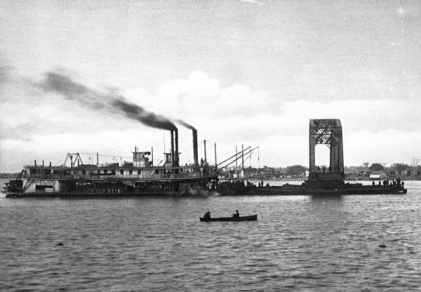 A floating span of Southern Pacific Railroad Bridge over the Atchatalaya River under construction. A steamboat is pulling the bridge section on a raft and a small boat with two passengers is in the foreground.