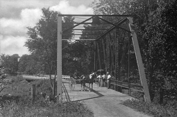 A man in a horse-drawn carriage and pedestrians with umbrellas relax on Coon Creek Bridge, a small steel bridge through the country.