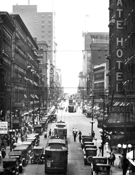 Elevated view of Main Street. City scene showing rows of parked cars, tall buildings, pedestrians, trolleys and advertising signs.
