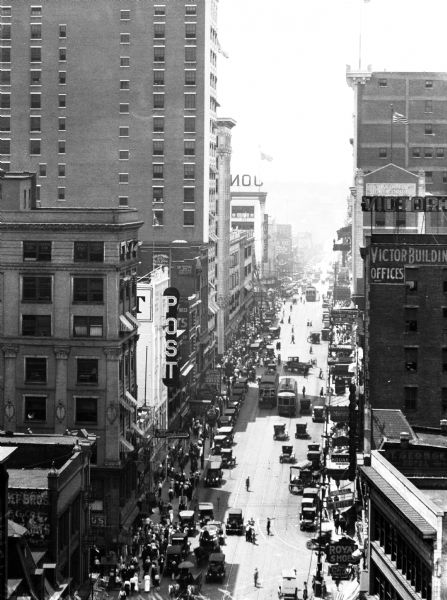 Elevated view of Main Street showing tall buildings, cars, pedestrians trolleys and advertising signs.
