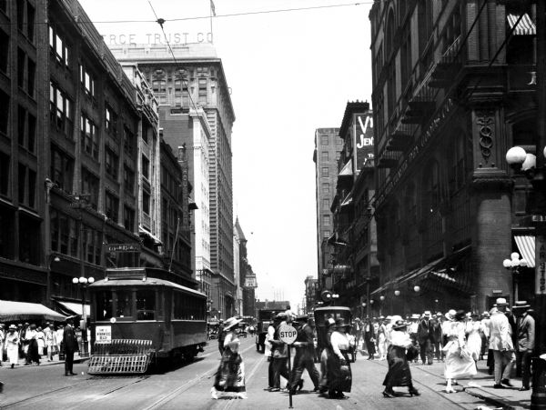 Street level view of Grand Avenue showing a trolley, a crosswalk, crowded sidewalks and tall buildings.