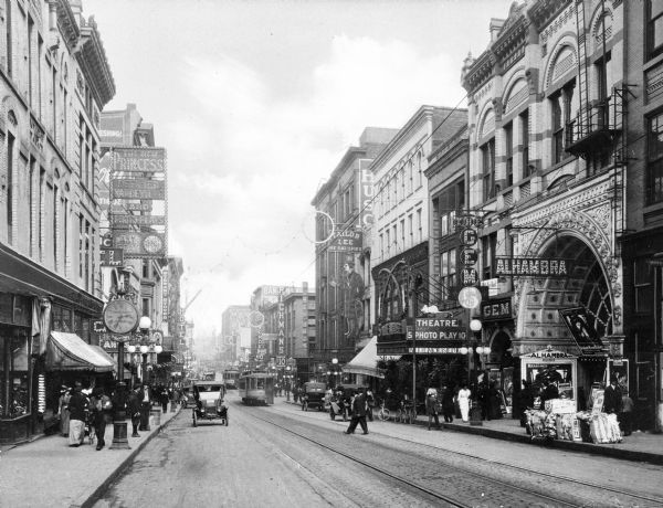 Street-level view of Seventh Street showing tall stone buildings, advertising signs, foot traffic, cars and a trolley.