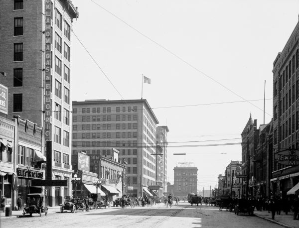 Street-level view of Broadway Avenue showing tall stone buildings, cars, carriages, horses and pedestrians in the distance.