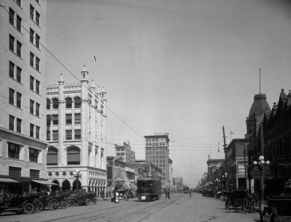A street scene showing tall stone buildings, parked cars and a trolley.