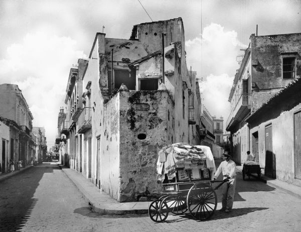 A street vendor with a cart on a narrow cobblestone street through a residential neighborhood of stucco houses in Havana, Cuba.