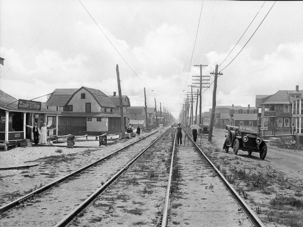 Railway tracks along a residential roadway.  A car is parked by the road and a few people stand around the area near a small store.