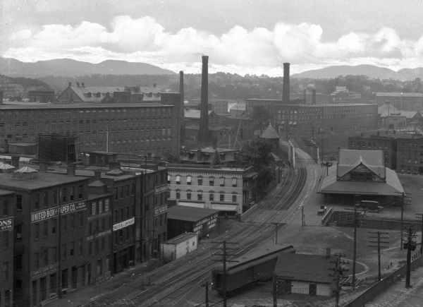 Elevated view of industrial section of town showing railroad tracks, brick warehouses, smokestacks and the mountains on the outskirts of town in the background.