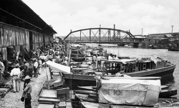 A view of small boats docked at a waterfront market in Tampico, Mexico showing a crowd of men in front of a waterfront warehouse, a steel bridge in the background and buildings on the opposite shore.