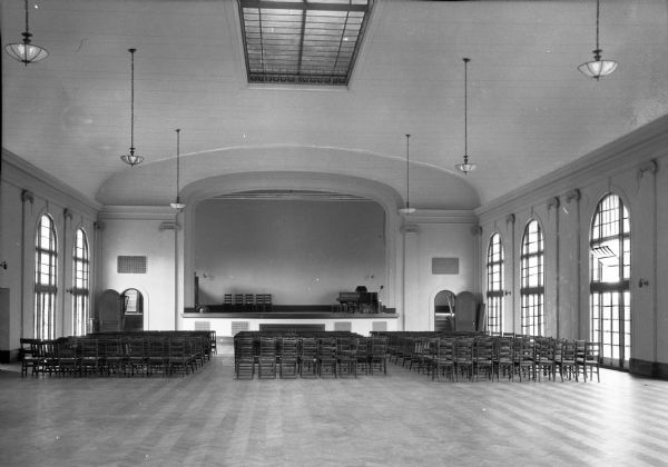 The Sea View Hospital auditorium interior, showing a stage, rows of wooden folding chairs and floor-to-ceiling windows.