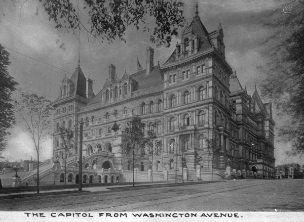 A view of the capitol building from Washington Avenue. Built in the neo-Renaissance style and architecturally modeled on the Hotel de Ville of Paris, France, the building has a stone causeway and many arched windows. Small trees frame the scene. Caption reads: "The Capitol  from Washington Avenue."