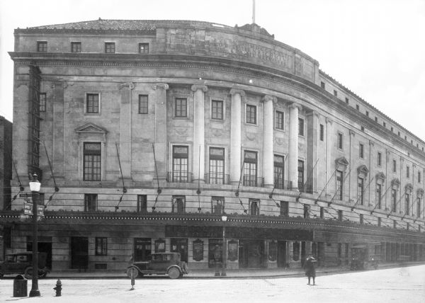 Front view of the Eastman Theatre, which opened in 1922, from across the street.  The large curving stone building with classical details wraps around the block and features decorative lighting.  A few cars are parked in front of the theatre and a pedestrian is crossing the street.