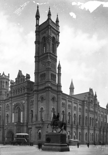 View of the stone Norman style Masonic Temple Grand Lodge and its tower.  A statue of General John Fulton Reynolds on horseback is in the square in front of the building and a few pedestrians and a double-decked horse-drawn carriage can also be seen near the building.
