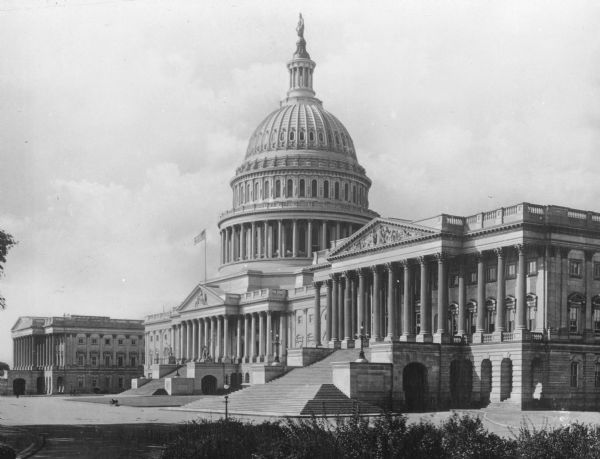A view of the Capitol Building from the East front showing extended stone stairs, exterior columns, bas-relief, the dome and the stone plaza in front.