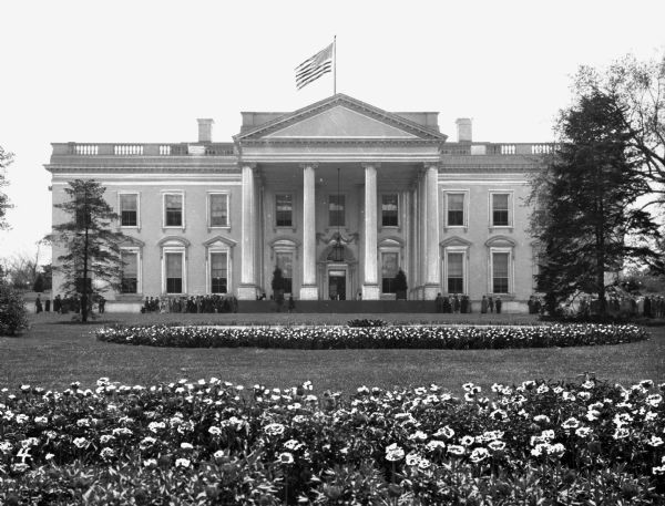 A view of the White House from the front lawn showing blooming flowers and small groups of people gathered by the drive.