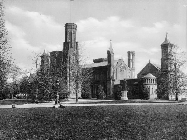 A view of the Smithsonian Institute's 'Castle' building from a nearby lawn.  The sprawling, crenelated, multi-part stone structure is bordered by some trees and bushes and a statue stands in front of one section of the building.