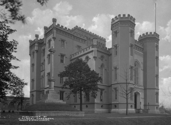 A view of the Old Louisiana State Capitol building from the front lawn.  The castle has crenelated battlements and a decorative stone post on the lawn in the foreground of the photo.  Published by J. Albert Anderson, Books and Stationary.