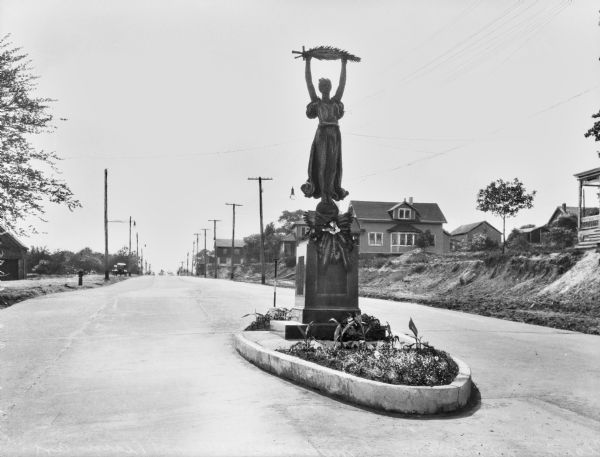 The Pleasant Plains Memorial statue honoring the soldiers from the fifth ward who fought in World War II.  The bronze statue of a woman holding a palm and a sword aloft stands in a narrow traffic island.  Houses along the road are visible in the background.