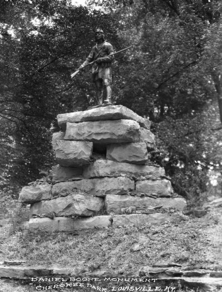 Low view of a bronze statue of Daniel Boone standing atop a pedestal of rough-hewn rocks. Leafy tree limbs form the backdrop.