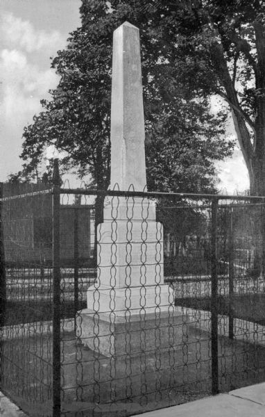 Monument to John Brown and the men killed during the Battle of Osawatomie. Those slain in the battle are buried underneath the monument. Chain link fence surrounds the small obelisk and a large tree's leafy cascade cuts across the background.