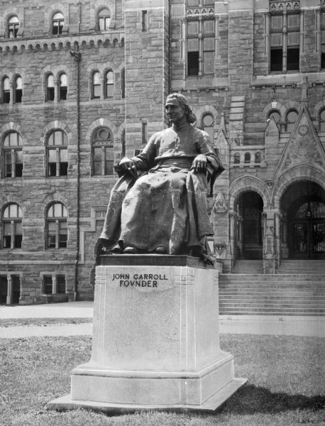 Seated statue of John Carrol, first Archbishop of Baltimore and founder of Georgetown University.  The sculpture has a small pedestal and part of the stone facade of Healy Hall forms the background of the photo.