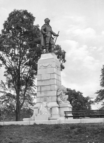 Statue dedicated to the French geographer, explorer and colonialist Samuel de Champlain (1567-1635).  A bronze man in a cape stands at the top of the pedestal and stone statues of a crouching Native American man and the stern of a canoe sit at the base of the pedestal.  Trees, a bench and grass of the surrounding park also appear in the photo.