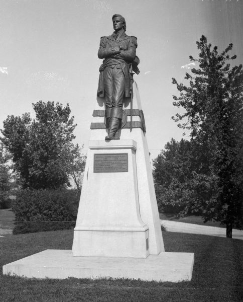 A bronze statue of Revolutionary War General, George Roberts Clark (1752-1818).  The statue leans against a two-tiered stone base and is framed by park greenery.