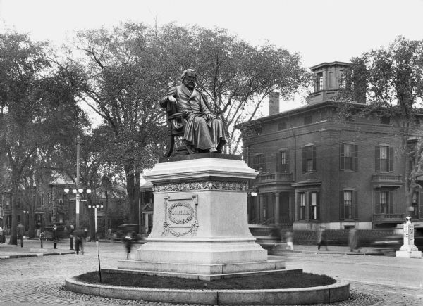 Close-up of the Henry Wadsworth Longfellow monument and the surrounding cobbled square.  Installed in 1888 and sculpted by Franklin Simmons, the monument features a bronze of Longfellow seated and an embellished stone pedestal.