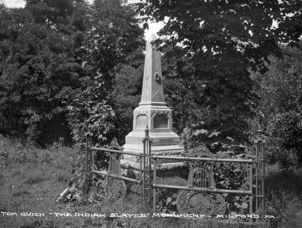 The grave marker of Tom Quick, Jr., who reputedly killed many Native Americans after he allegedly witnessed his father killed by a group of Native Americans. The small stone obelisk stands behind an ornate fence among a stand of trees. Caption reads: "Tom Quick 'The Indian Slayer' Monument." The monument was removed after being vandalized in 1997. 
