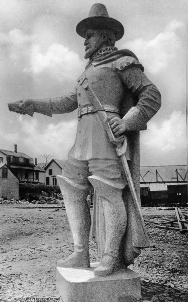 Close-up of a statue of Captain Myles Standish, a military leader of the Plymouth Colony.  Begun in 1892, the statue depicts Standish grasping his sword in its scabbard at his side and gesturing with his other hand.  Large wooden buildings are visible across a barren square in the background.