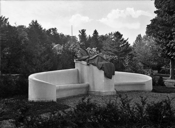 A statue of Dr. Edward Livingston Trudeau, a tuberculosis specialist, on the grounds of the Trudeau Sanitorium Historic District. Installed in 1918, the bronze Gutzon Borglum statue depicts Trudeau reclining and wrapped in a blanket atop a stone pedestal connected to curving stone benches. A gravel path leads to the monument through wooded grounds.