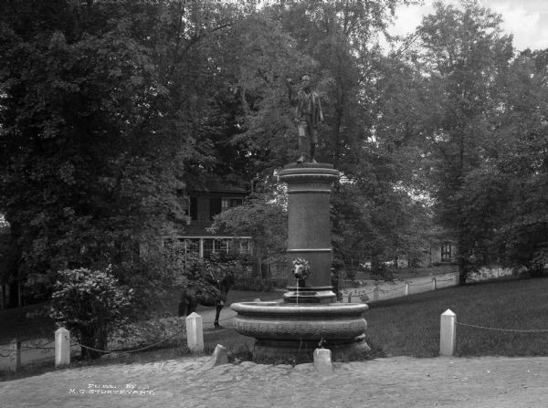 View of a statue and fountain in a grassy park depicting a boy selling a newspaper.  Installed in 1895 and funded by Colonel William L. Brown, the statue celebrates the newspaper industry.  A boy on a horse looks at the statue from just outside of the park and trees shade the adjacent neighborhood.
