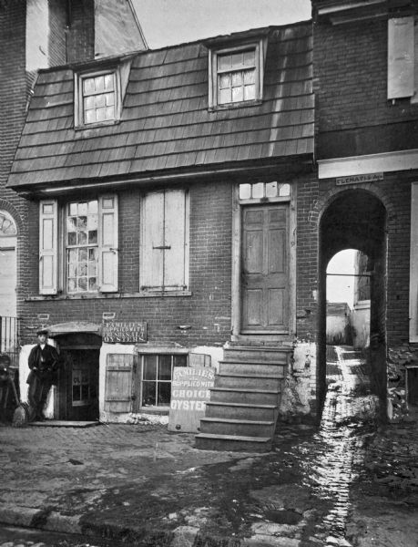 A city view of a brick building and a cobbled alleyway called Clements Avenue. Two individuals are standing outside the entrance of the building, which bears signs advertising fresh and salted oysters.