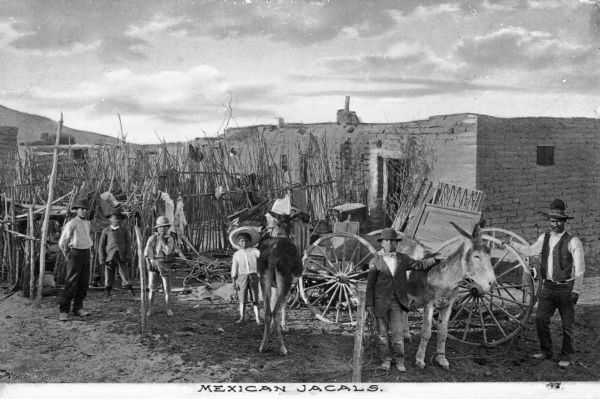 A Mexican Jacal (an adobe style housing structure) with five boys and one man standing in front with two donkeys and a cart.